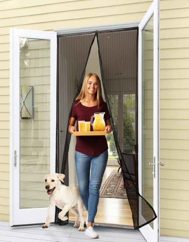 A person is standing in a doorway with a mesh black trim door curtain, holding a tray with a beverage, accompanied by a dog. The door curtain is designed to keep insects out and is hands-free with a magnetic closure.